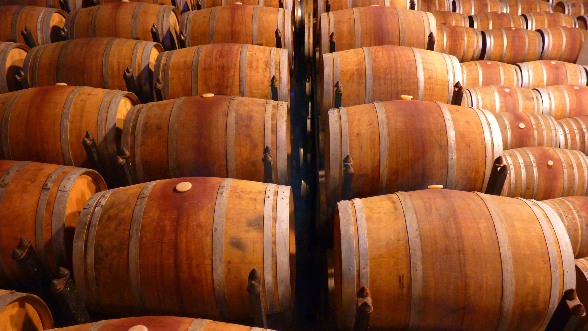 Rows of wooden wine barrels stacked in a cellar or warehouse, showing traditional oak aging barrels with metal hoops used for wine or spirits production